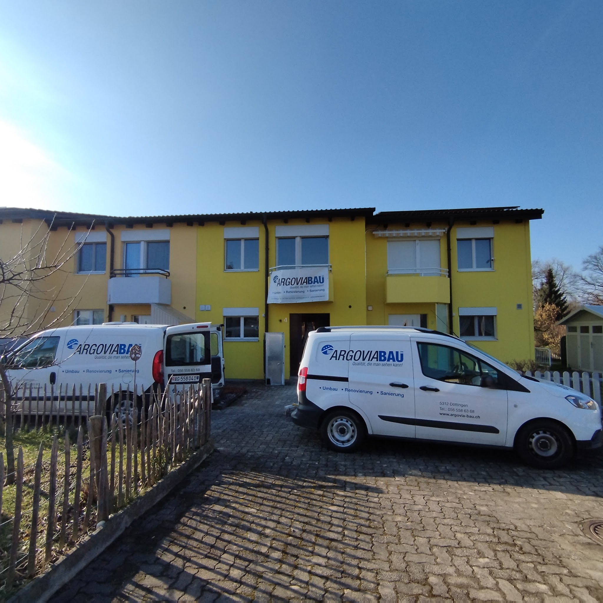 Gelbes Mehrfamilienhaus mit Firmenbanner und zwei Argovia-Bau-Transportern